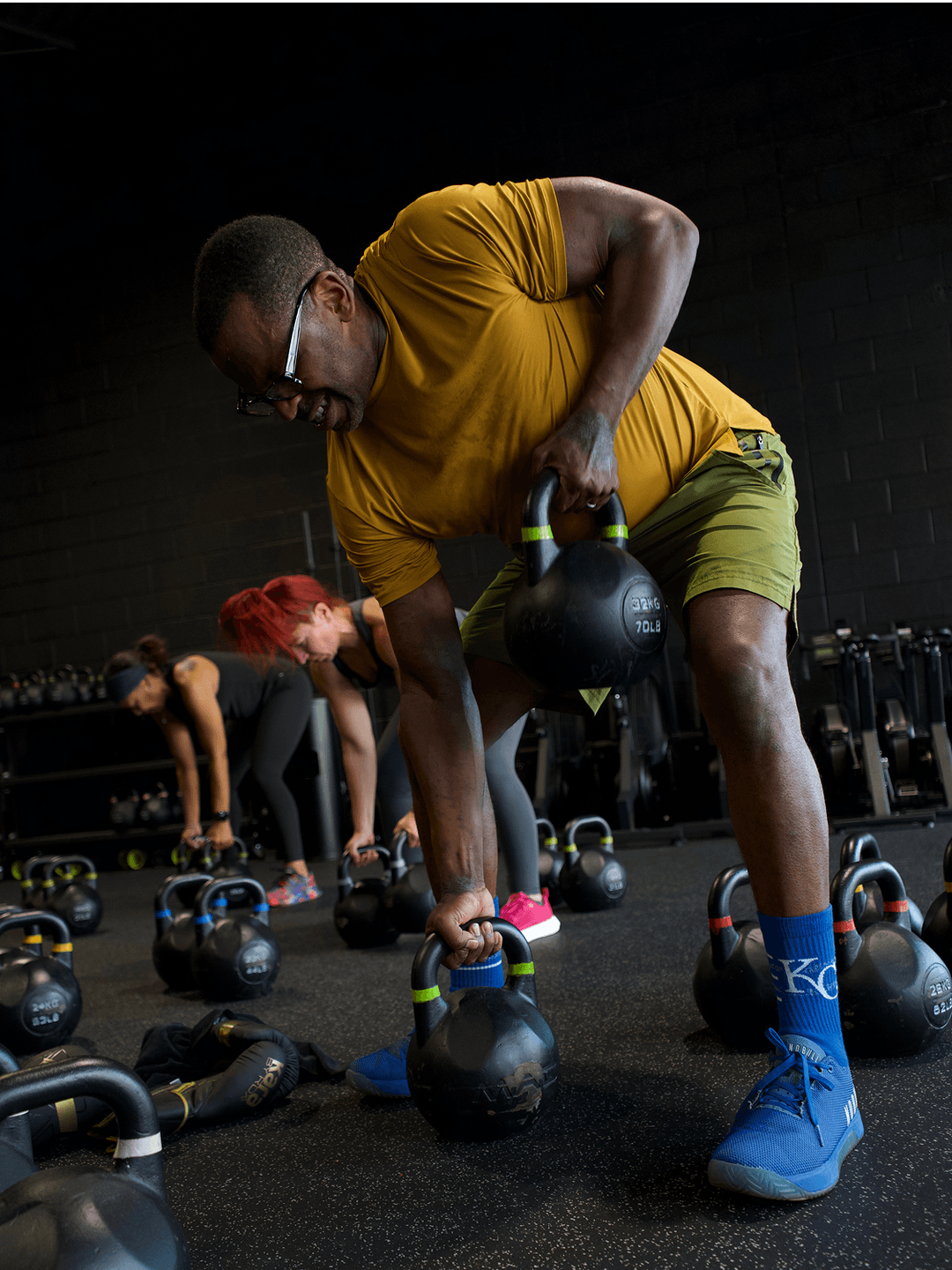A member bends to lift a kettlebell on the studio floor, with racks of kettlebells and other members behind.