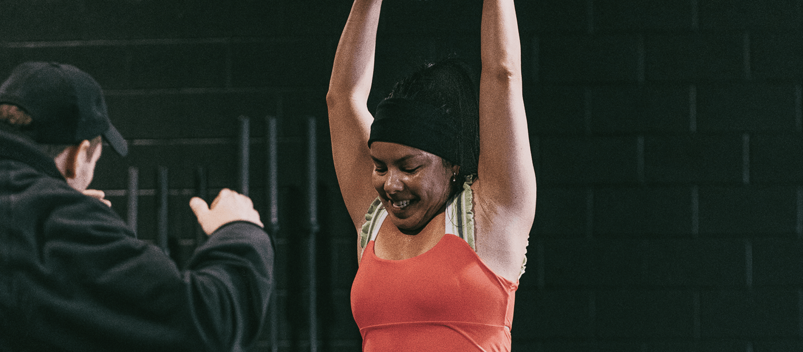 A member raises her arms overhead in the studio while a person in a cap stands beside her.