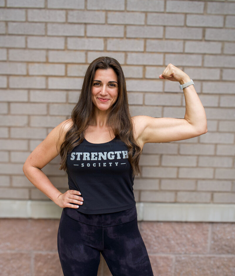 Coach Katie smiling and flexing in front of a brick wall in a Strength Society tank top.